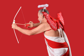 Naklejka premium Mature man dressed as Cupid shooting bow on red background. Valentine's Day celebration