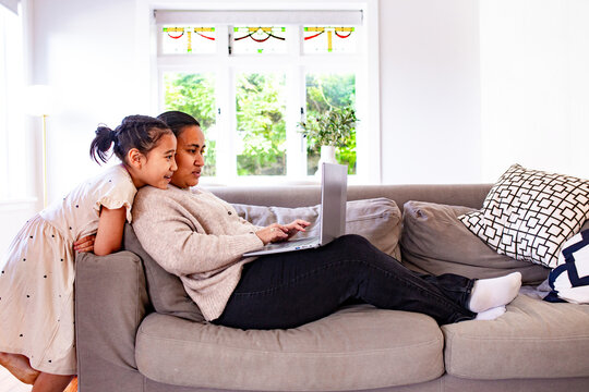Young Samoan girl standing behind mum lying on the couch working on her laptop