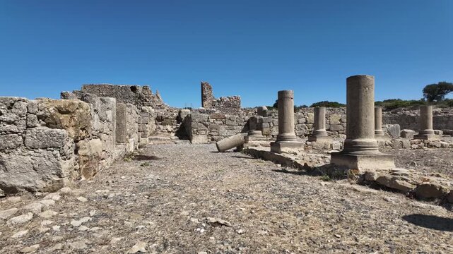 Stabilized handheld shot walking through the Roman ruins of King Juba II's palace in Lixus. The footage features ancient stone columns and walls under a blue sky in Larache, Morocco.