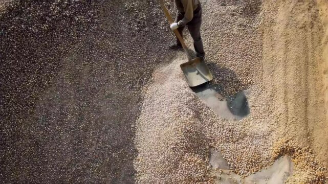 Worker moves gravel with a shovel at a construction site in the afternoon sun on a clear day, focusing on the task at hand with careful attention and effort