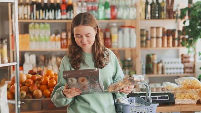Adolescence girlie examines packaged sweet products in store. Market visitor chooses cookies. She stands in sales area of grocery and thinks about buying desserts pastry