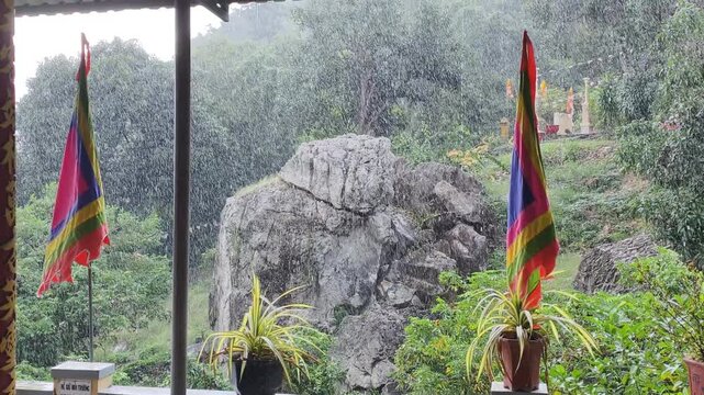 Wide view shows Tropical Rain with heavy water drops falling over mountain pagoda. Colorful Buddhist flags wave during Tropical Rain amidst misty rocks and green jungle.