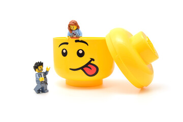 Naklejka premium Lego minifigures are at the large yellow head with funny face is sticking out the tongue isolated on white. Editorial illustrative image of popular plastic toy.