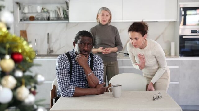 Upset middle-aged man sitting at table while two women quarreling to him standing behind in the kitchen decorated with Xmas-tree. High quality 4k footage