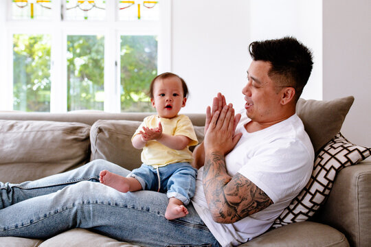 Asian father and baby girl clapping while seated on the couch