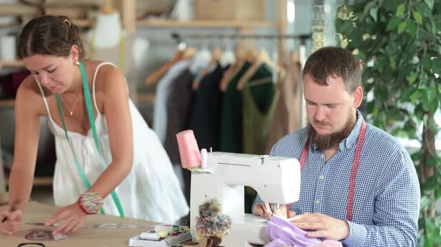 Focused male tailor sewing pastel fabric at machine with precision, while young female colleague sketching patterns on kraft paper nearby in shared studio