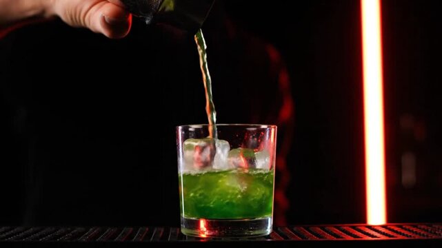 Professional Bartender Pouring a Vibrant Green Cocktail from a Shaker into a Glass filled with Ice Cubes in a Modern Dark Bar Setting.