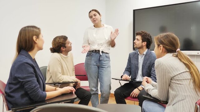Young female business coach explaining topic while standing against group of people sitting at desks in front of her