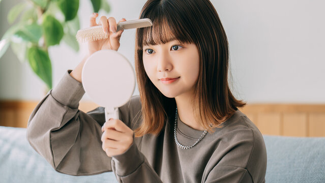 鏡を見ながら前髪を整える日本人女性,室内でのヘアケアと身だしなみ, Japanese woman adjusting her bangs while looking in a mirror during indoor hair care