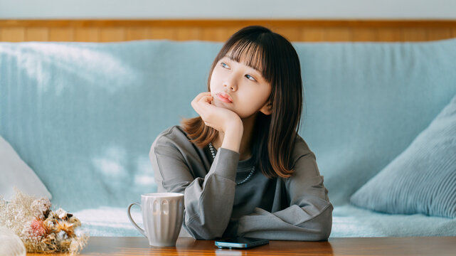 テーブルで頬杖をつき考え事をする日本人女性,コーヒーとスマートフォンのある室内風景, Japanese woman resting her chin and thinking at a table with coffee and a smartphone indoors