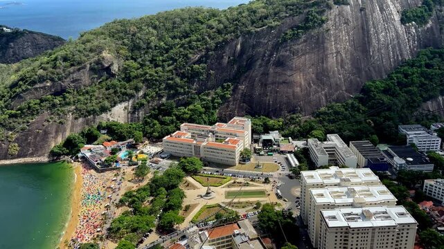 A sweeping pan-right view from Morro da Urca showing the crowded Praia Vermelha beach, the historic IME building, and the lush Atlantic Forest surrounding the granite peaks of Rio.