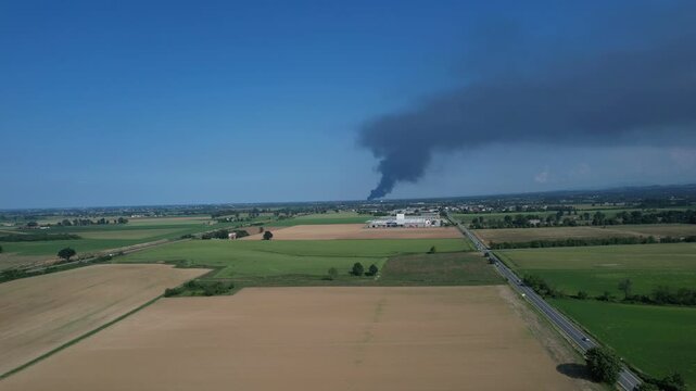 Drone view of dense black smoke rising from a burning plastic factory and drifting over farmland highlighting air pollution, dioxin contamination risk and an active environmental emergency.