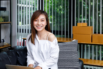 Portrait of beautiful young Asian woman smiling at camera while working on laptop at home. SME online seller concept with parcel boxes in background.