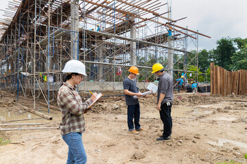 Asian female engineer writing on clipboard checklist while male colleagues discuss in background. Quality control manager monitoring building project progress at construction site.