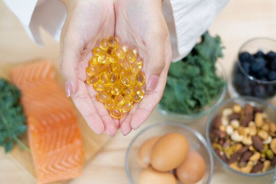 Hands holding fish oil capsules above salmon, eggs, nuts, vegetables, and blueberries on a table, showing nutrition, dietary supplement, and healthy lifestyle concept.