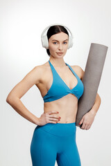 Fit woman in blue sportswear wearing headphones holds a yoga mat with a confident pose against a plain white background, showcasing athleticism and fitness lifestyle