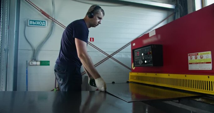 Machine operator feeding sheet metal into industrial guillotine shear inside factory workshop. Blue-collar manufacturing job focused on metal cutting and production workflow