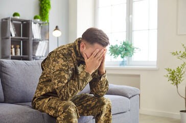 Fototapeta premium Man in military camouflage uniform sits on couch expressing deep sadness or emotional stress, suffers from post-traumatic stress disorder, need psychological aid. Mental health, counselling