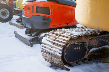 Snow covered steel track on compact excavator in winter equipment yard, with orange machine body and dozer blade blurred behind, heavy construction machinery stored outdoors in cold season © K