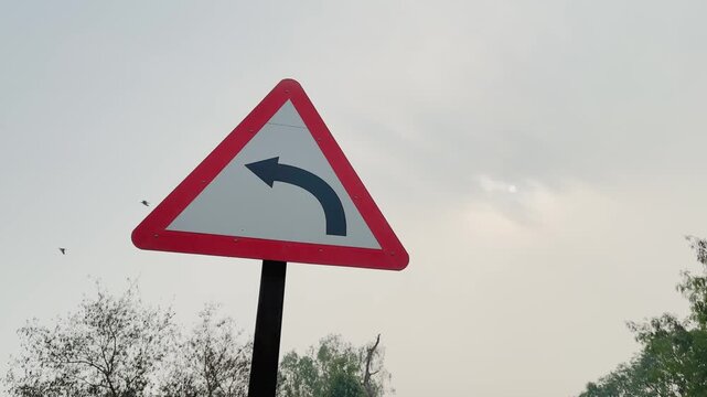 Camera orbiting around a triangular left turn warning road sign on a pole, red border and black arrow visible against pale sky with distant trees in daylight conditions.