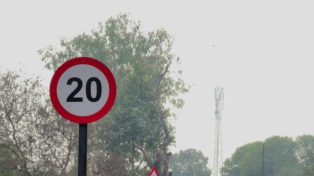 Static shot of a circular speed limit 20 road sign mounted on a pole, red border and bold numerals visible with trees and distant tower against hazy sky.