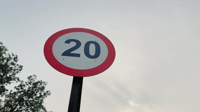 Camera orbiting around a circular speed limit 20 road sign mounted on a pole, red border and bold numerals visible against pale sky and trees in daylight.