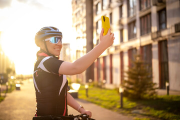 Smiling woman cyclist taking selfie with yellow smartphone against city buildings. Joy of movement and social sharing in urban lifestyle.