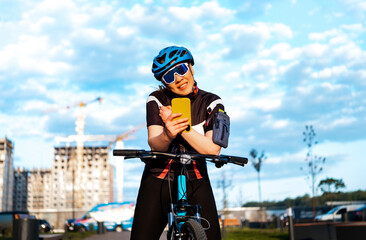Woman cyclist leaning on bicycle handlebars and using smartphone in new residential district. Modern urban infrastructure for active recreation.