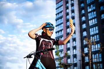 Happy woman cyclist in helmet holding smartphone and celebrating ride on city background. Digital lifestyle and outdoor cardio training.