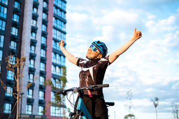 Woman cyclist celebrating successful training with thumbs up and raised arms against blue sky. Inspiration and pleasure from sports activity.