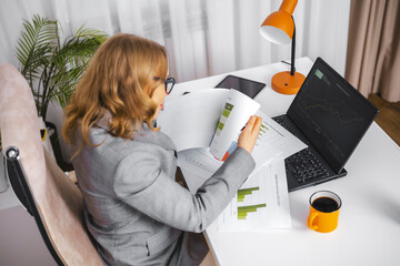 Professional woman wearing glasses analyzing financial charts and marketing reports at white office desk. Data-driven decision making, business planning, financial expertise, corporate analytics.