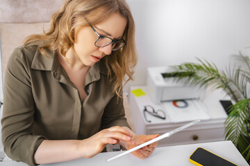 Surprised woman in glasses using digital tablet and browsing data at office desk with printer. Rapid technology adoption, digital transformation, shock from news, modern software usage.
