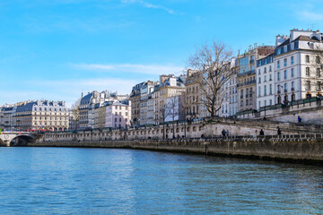 Fototapeta premium Calm river scene in Paris with classic buildings along the Seine, elegant facades, mansard roofs and a stone bridge on the left. Clear blue sky and bare trees create a peaceful early spring atmosphere