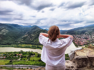 Naklejka premium Woman looks out over valley with river and mountains while standing on a rocky ledge in a cloudy landscape