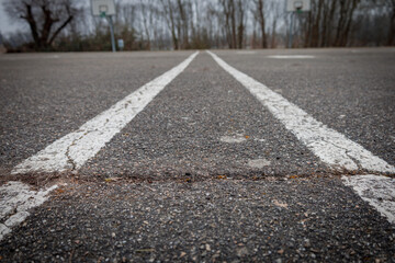 Obraz premium Faded white lines stretch across a weathered asphalt court, leading towards distant, blurry basketball hoops amidst bare trees. The scene evokes a sense of quiet abandonment and past games.