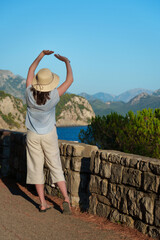 Back view of a young woman in a straw hat raising her hands towards the sky, enjoying the majestic mountain and sea landscape during sunset.
