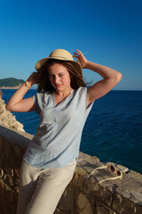 Pretty young woman with a hat posing by the sea at sunset, summer vacation travel in Balkans.