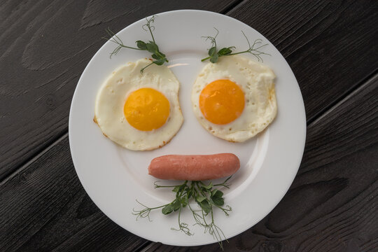 Two fried eggs with a smiling sausage in between on a white plate, dark wooden background.