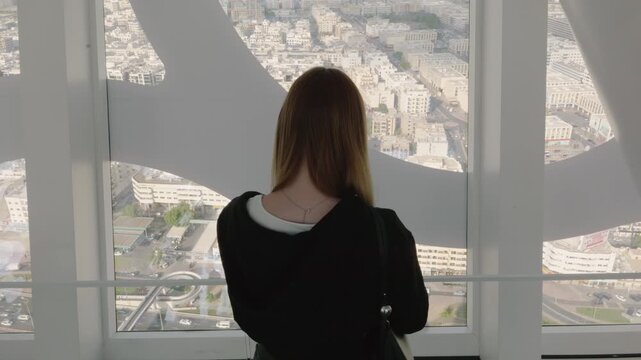 Young caucasian woman standing indoors and looking at city skyline through panoramic window at modern observation deck, representing sightseeing and urban travel experience.