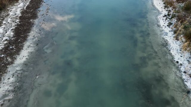 Aerial drone shot of a narrow turquoise river flowing through a snowy shoreline with dry yellow reeds and ice patches on a cold day