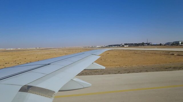 POV from an airplane window over the wing, taxiing and taking off at Queen Alia International Airport, Jordan, on a bright, sunny day with terminal buildings visible
