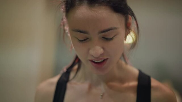 asian woman close up after workout, flushed cheeks, sweat on skin, soft smile, focused gaze, hair tied in ponytail, sports bra straps, studio bokeh lights, trainer blurred in background, candid