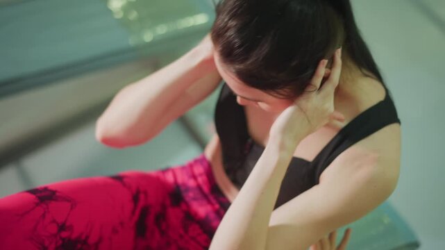 woman doing ab crunches on bench with trainer assisting legs, wearing black sports bra and red patterned leggings, hands at head, close up of core contraction, gym bench and equipment in background,