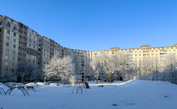 A winter city courtyard with gray multi-story panel buildings (Khrushchev-era or Brezhnev-era buildings) covered in a thin layer of snow and frost, under a bright blue sky