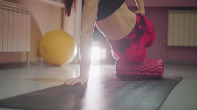 Woman on mat using foam roller to release glute and hip tension, soft morning light, intimate studio, yoga ball in background, slow controlled movements, calming recovery atmosphere, focused breathing
