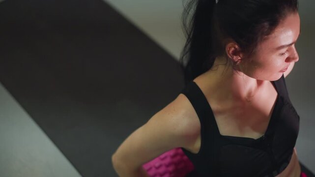 Asian woman on mat foam roller for recovery, massaging lower back and hamstrings with slow controlled motion, soft studio lighting, black sports bra, pink textured roller, focused breathing, calm mood