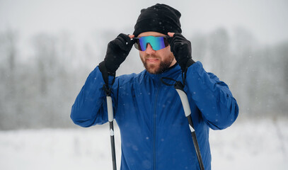 Male skier wearing blue jacket and black beanie adjusts colorful sunglasses while holding ski poles in snowy landscape with blurred trees in the background