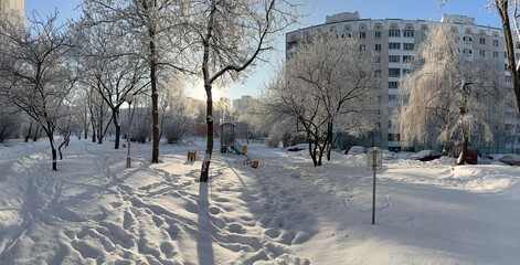 Obraz premium A winter city courtyard with gray multi-story panel buildings (Khrushchev-era or Brezhnev-era buildings) covered in a thin layer of snow and frost, under a bright blue sky