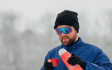 Male skier wearing blue jacket and sunglasses holds red cup and thermos while standing in snowy landscape with falling snowflakes in winter season