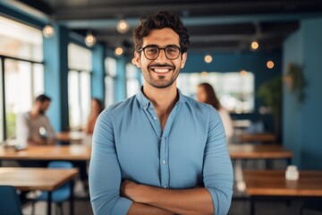 Obraz premium Young professional man with glasses standing with arms crossed, smiling at the camera in a contemporary workspace